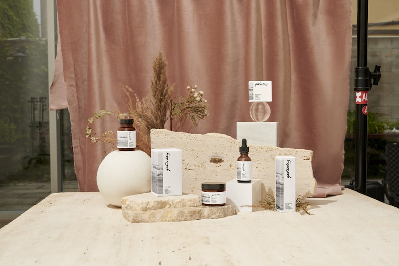 A collection of skincare products displayed on a light beige stone surface. The background features a soft, muted pink curtain with dried floral arrangements, adding an organic and natural element. The products are arranged in an aesthetically pleasing manner, with variations in height and texture for visual interest.