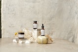 A minimalist arrangement featuring skincare products including a jar, a bottle with a dropper, and another jar. These items are placed on white blocks, accompanied by delicate dried flowers and a decorative stone object. The background is a textured, light-colored surface that enhances the serene and natural aesthetic.