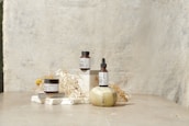 A minimalist arrangement featuring skincare products including a jar, a bottle with a dropper, and another jar. These items are placed on white blocks, accompanied by delicate dried flowers and a decorative stone object. The background is a textured, light-colored surface that enhances the serene and natural aesthetic.