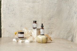A minimalist arrangement featuring skincare products including a jar, a bottle with a dropper, and another jar. These items are placed on white blocks, accompanied by delicate dried flowers and a decorative stone object. The background is a textured, light-colored surface that enhances the serene and natural aesthetic.