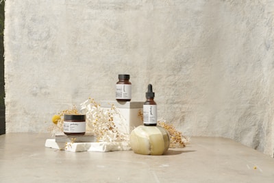 A minimalist arrangement featuring skincare products including a jar, a bottle with a dropper, and another jar. These items are placed on white blocks, accompanied by delicate dried flowers and a decorative stone object. The background is a textured, light-colored surface that enhances the serene and natural aesthetic.