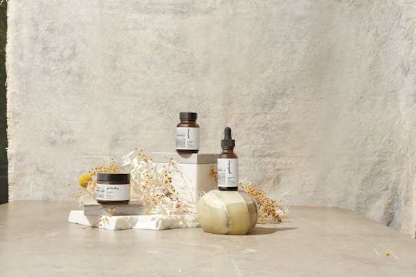 A minimalist arrangement featuring skincare products including a jar, a bottle with a dropper, and another jar. These items are placed on white blocks, accompanied by delicate dried flowers and a decorative stone object. The background is a textured, light-colored surface that enhances the serene and natural aesthetic.
