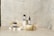 A minimalist arrangement featuring skincare products including a jar, a bottle with a dropper, and another jar. These items are placed on white blocks, accompanied by delicate dried flowers and a decorative stone object. The background is a textured, light-colored surface that enhances the serene and natural aesthetic.