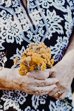 An elderly person with detailed, patterned clothing, holding a small bouquet of yellow flowers in their hands, adorned with jewelry such as rings.
