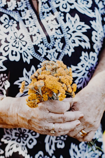 An elderly person with detailed, patterned clothing, holding a small bouquet of yellow flowers in their hands, adorned with jewelry such as rings.