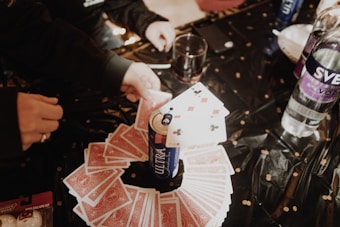 A table with a black tablecloth is laid out with playing cards arranged in a circular pattern. A can of Michelob Ultra stands in the center with a few playing cards on top of it. Various hands are seen reaching for the cards. Other items on the table include a bottle of Svedka vodka, a drink in a glass, and a box of Buffalo Wild Wings Blazin' Reaper wing sauce gift set.