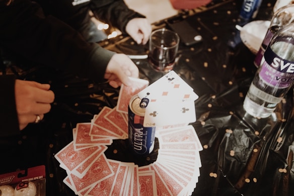 A table with a black tablecloth is laid out with playing cards arranged in a circular pattern. A can of Michelob Ultra stands in the center with a few playing cards on top of it. Various hands are seen reaching for the cards. Other items on the table include a bottle of Svedka vodka, a drink in a glass, and a box of Buffalo Wild Wings Blazin' Reaper wing sauce gift set.