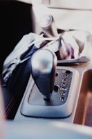 A close-up view of a car's gear shift panel with an automatic transmission. The panel displays various gear options including P, R, N, D3, 2, and L. The gear lever is prominently visible in the foreground, surrounded by some interior trim. The lighting suggests it is daytime, possibly with sunlight filtering into the car.