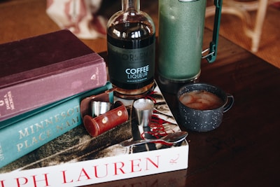 A cozy setup on a wooden table features three books stacked in the background, a bottle of coffee liqueur, a green thermos, a dark speckled coffee mug with a hot beverage, and a red case with metal shot glasses alongside a spoon.