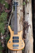 A wooden electric bass guitar with four strings is placed on a bed of organic materials, including moss-covered and bark-stripped logs. Surrounding the guitar, there are green leaves, twigs, and some dried leaves, indicating an outdoor, natural setting.