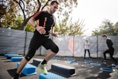 A man is actively participating in an outdoor fitness session, jumping over blue platforms with agility. Two other people in the background are also engaging in exercise, with rings scattered on the ground. The area is fenced and surrounded by trees, suggesting a park or recreational setting.
