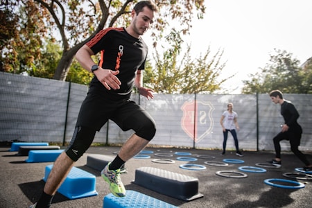 A man is actively participating in an outdoor fitness session, jumping over blue platforms with agility. Two other people in the background are also engaging in exercise, with rings scattered on the ground. The area is fenced and surrounded by trees, suggesting a park or recreational setting.