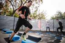 A man is actively participating in an outdoor fitness session, jumping over blue platforms with agility. Two other people in the background are also engaging in exercise, with rings scattered on the ground. The area is fenced and surrounded by trees, suggesting a park or recreational setting.