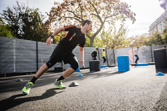 Several people engage in an outdoor fitness training session. One individual in the foreground is running between cones and obstacles. Trees with autumn leaves and a tall building are visible in the background, and there are cones and cylindrical barriers scattered around.