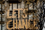 Large golden letters spell out 'LET'S CHANGE' displayed on the exterior of a building with tall windows and decorative columns. Skeletal branches with sparse yellow leaves partially obscure the text.