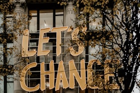 Large golden letters spell out 'LET'S CHANGE' displayed on the exterior of a building with tall windows and decorative columns. Skeletal branches with sparse yellow leaves partially obscure the text.