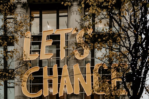 Large golden letters spell out 'LET'S CHANGE' displayed on the exterior of a building with tall windows and decorative columns. Skeletal branches with sparse yellow leaves partially obscure the text.