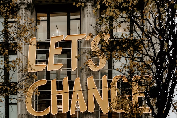 Large golden letters spell out 'LET'S CHANGE' displayed on the exterior of a building with tall windows and decorative columns. Skeletal branches with sparse yellow leaves partially obscure the text.