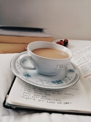 A white teacup filled with light brown tea rests on an open notebook. The cup is placed on a matching saucer with a floral pattern. Beside the notebook are stacked books, and dried flowers are seen in the background. The notebook has a handwritten to-do list with neat bullet points.