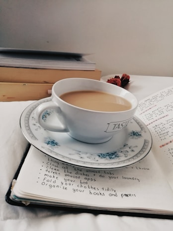 A white teacup filled with light brown tea rests on an open notebook. The cup is placed on a matching saucer with a floral pattern. Beside the notebook are stacked books, and dried flowers are seen in the background. The notebook has a handwritten to-do list with neat bullet points.