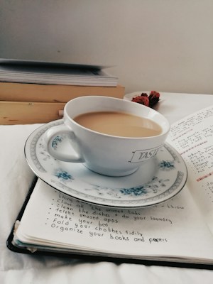 A white teacup filled with light brown tea rests on an open notebook. The cup is placed on a matching saucer with a floral pattern. Beside the notebook are stacked books, and dried flowers are seen in the background. The notebook has a handwritten to-do list with neat bullet points.
