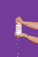 Two hands hold a can labeled 'She Can Coastal Berry' with liquid dripping from it, set against a vibrant purple background. The hands are positioned as though opening the can, suggesting action and refreshment.