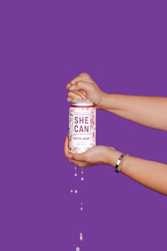 Two hands hold a can labeled 'She Can Coastal Berry' with liquid dripping from it, set against a vibrant purple background. The hands are positioned as though opening the can, suggesting action and refreshment.