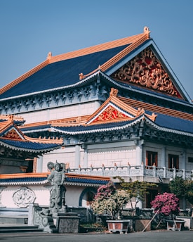 A traditional Asian temple with intricate architectural details, featuring colorful roofs with ornate patterns in red and gold. A stone statue is positioned at the forefront, surrounded by potted plants and trees. The blue sky creates a serene backdrop.