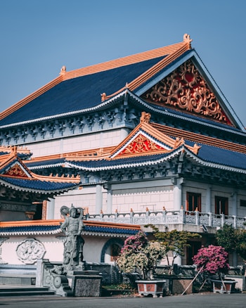 A traditional Asian temple with intricate architectural details, featuring colorful roofs with ornate patterns in red and gold. A stone statue is positioned at the forefront, surrounded by potted plants and trees. The blue sky creates a serene backdrop.