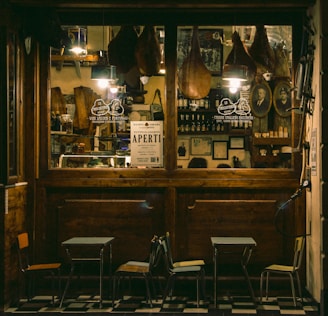 A cozy Italian deli or cafe with a wooden exterior, displaying hanging cured meats, framed pictures, and shelves stocked with bottles and jars. A sign reading 'Aperti' indicates it is open. Outside are small tables and chairs on a tiled floor.