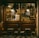 A cozy Italian deli or cafe with a wooden exterior, displaying hanging cured meats, framed pictures, and shelves stocked with bottles and jars. A sign reading 'Aperti' indicates it is open. Outside are small tables and chairs on a tiled floor.