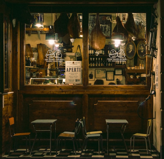 A cozy Italian deli or cafe with a wooden exterior, displaying hanging cured meats, framed pictures, and shelves stocked with bottles and jars. A sign reading 'Aperti' indicates it is open. Outside are small tables and chairs on a tiled floor.