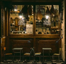 A cozy Italian deli or cafe with a wooden exterior, displaying hanging cured meats, framed pictures, and shelves stocked with bottles and jars. A sign reading 'Aperti' indicates it is open. Outside are small tables and chairs on a tiled floor.