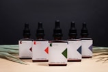 A row of five brown glass dropper bottles, each labeled with 'Batch' and featuring different geometric designs. The labels indicate various CBD concentrations with product names like 'Original' and 'Calm'. The background is a dark shade, while dried palm leaves are arranged at the base.