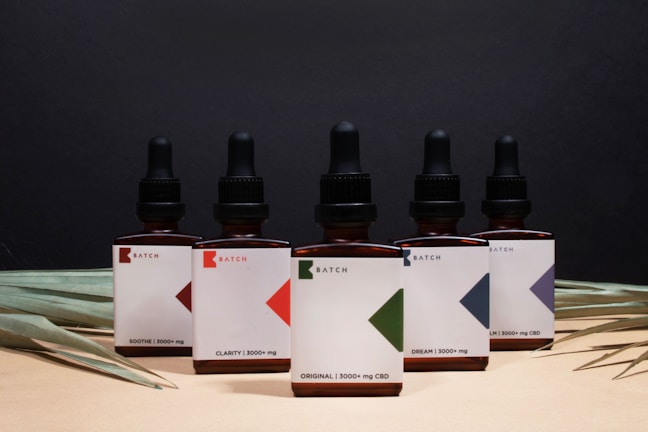 A row of five brown glass dropper bottles, each labeled with 'Batch' and featuring different geometric designs. The labels indicate various CBD concentrations with product names like 'Original' and 'Calm'. The background is a dark shade, while dried palm leaves are arranged at the base.