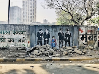 A vibrant mural is painted on a worn concrete wall, depicting a group of four people dressed in black suits and ties, with a young girl in a blue dress standing among them. The wall contains text in a regional script, possibly Hindi or Marathi, and is adorned with artistic elements such as flowers. In front of the mural, there is a pile of rubble consisting of rocks and broken concrete slabs. Behind the wall, modern high-rise buildings and leafless trees can be seen, suggesting an urban setting.