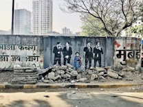 A vibrant mural is painted on a worn concrete wall, depicting a group of four people dressed in black suits and ties, with a young girl in a blue dress standing among them. The wall contains text in a regional script, possibly Hindi or Marathi, and is adorned with artistic elements such as flowers. In front of the mural, there is a pile of rubble consisting of rocks and broken concrete slabs. Behind the wall, modern high-rise buildings and leafless trees can be seen, suggesting an urban setting.