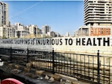 A large, bold message on a concrete wall reads 'Warning: Shopping is Injurious to Health.' In the background, there are several tall, modern apartment buildings. The foreground features a black metal fence and a road with some debris.
