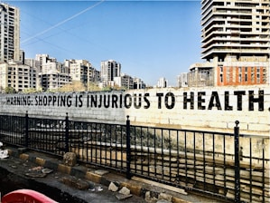 A large, bold message on a concrete wall reads 'Warning: Shopping is Injurious to Health.' In the background, there are several tall, modern apartment buildings. The foreground features a black metal fence and a road with some debris.