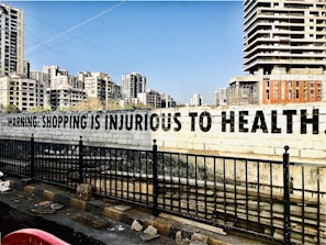 A large, bold message on a concrete wall reads 'Warning: Shopping is Injurious to Health.' In the background, there are several tall, modern apartment buildings. The foreground features a black metal fence and a road with some debris.