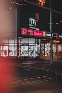 A nighttime urban scene featuring the exterior of a modern shopping center. Bright, colorful signs, including those of popular retail and cinema brands, illuminate the street. The entrance is decorated with festive lights, and a few people are visible, some inside the glass-walled building and others on the sidewalk. Streetcar tracks are visible on the ground.