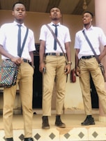 Three young men are standing on steps outside a building, dressed in matching uniforms consisting of white shirts, black ties, and khaki pants. One man holds a colorful bag, another has a phone in hand, and the third is wearing earphones. The mood is casual and confident.