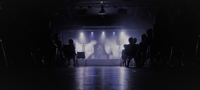 A dimly lit theater with an audience seated in chairs, facing a stage where performers are illuminated by spotlights. The background is dark, creating a contrast with the bright stage lights.