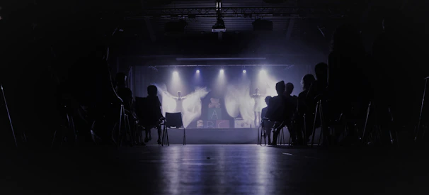 A dimly lit theater with an audience seated in chairs, facing a stage where performers are illuminated by spotlights. The background is dark, creating a contrast with the bright stage lights.