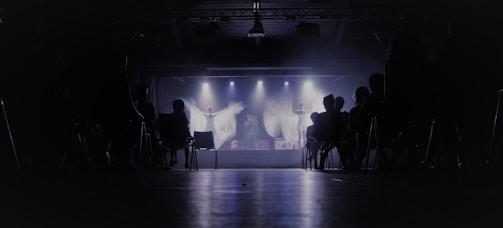 A dimly lit theater with an audience seated in chairs, facing a stage where performers are illuminated by spotlights. The background is dark, creating a contrast with the bright stage lights.