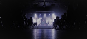 A dimly lit theater with an audience seated in chairs, facing a stage where performers are illuminated by spotlights. The background is dark, creating a contrast with the bright stage lights.
