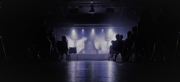 A dimly lit theater with an audience seated in chairs, facing a stage where performers are illuminated by spotlights. The background is dark, creating a contrast with the bright stage lights.