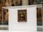 A famous framed painting displayed in a transparent glass case, viewed inside a museum with other artworks visible in the background. The painting is hung on a plain wall within the case.