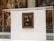 A famous framed painting displayed in a transparent glass case, viewed inside a museum with other artworks visible in the background. The painting is hung on a plain wall within the case.