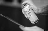 A hand is holding and using an aerosol spray can, applying it to an unseen surface. The can has visible branding and text.